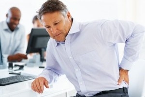 A man in discomfort sitting at a table holds his lower back